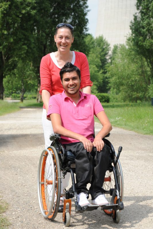 Jeune homme en fauteuil roulant aidé par une assistante de vie à ce déplacé dans un parc.