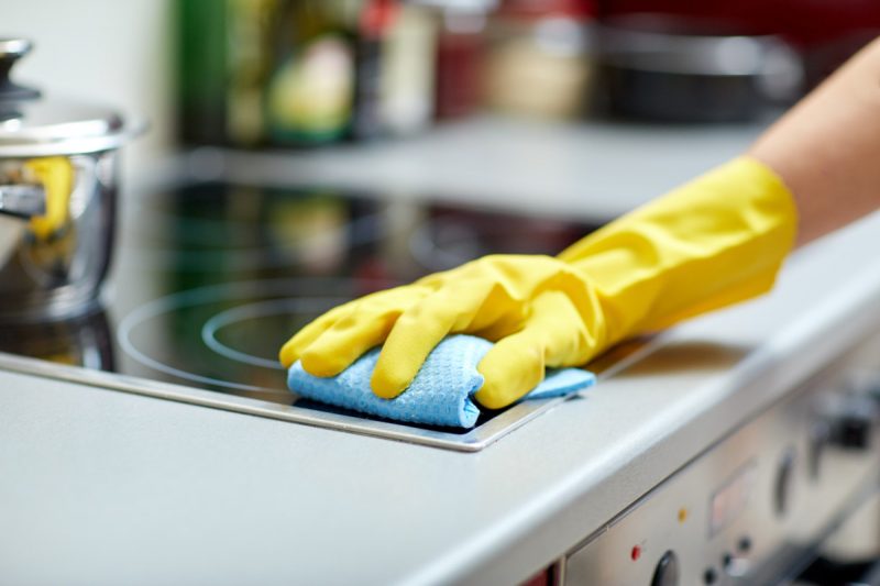 Détail sur la main d'un personne néttoyant une plaque à induction, la main est protégé par un lage gant jaune et tient un chiffon bleu.