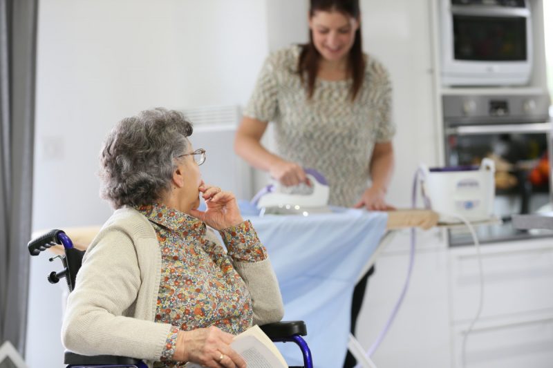 Jeune femme repassant des vêtements pour le compte d'une vielle dame en fauteuil roulant chez elle.