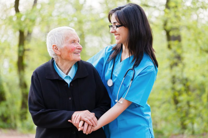 Viel homme aidé marché dans un parc par une femme, elle porte une tenue bleu ciel et un Stéthoscope autour du cou. ils sont vus de face