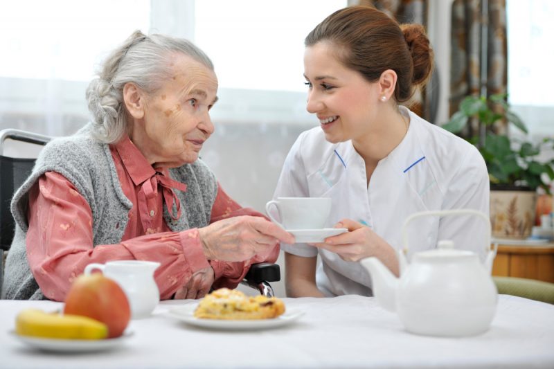 Deux femmes, celle de gauche très agée discute autour d'une table, celle de droite, en tenue d’infirmière lui tend une tasse.